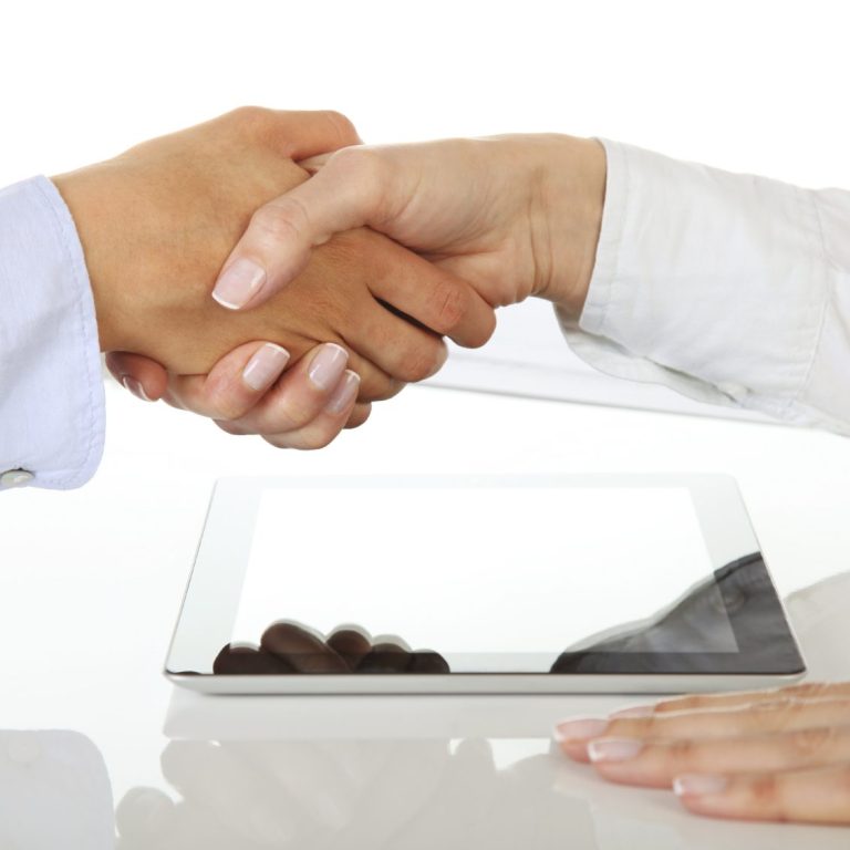 Dos manos se estrechan sobre una mesa, junto a una tableta y algunos chocolates.